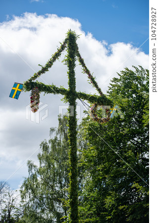 A pole and flag against green trees and blue sky. A maypole decorated, covered in flowers and leaves. 104728927