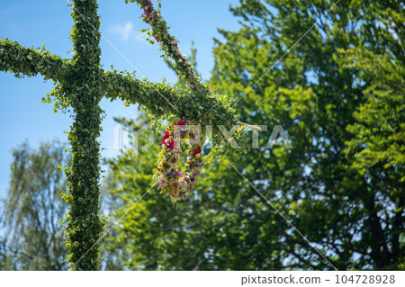 A pole and flag against green trees and blue sky. A maypole decorated, covered in flowers and leaves. 104728928