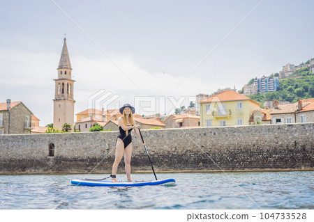 Young women Having Fun Stand Up Paddling in blue water sea in Montenegro. Against the backdrop of the Old Town of Budva. SUP Young women Having Fun Stand Up Paddling in blue water sea in Montenegro. Against the backdrop of the Old Town of Budva. SUP 104733528