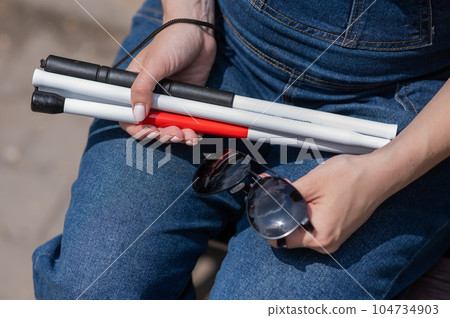 A blind woman sits on a bench and holds a folding tactile cane and sunglasses in her hands. 104734903