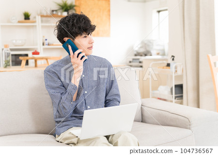Young man sitting on a sofa with a computer on his lap and calling on a smartphone 104736567