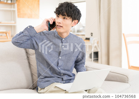 Young man sitting on a sofa with a computer on his lap and calling on a smartphone 104736568
