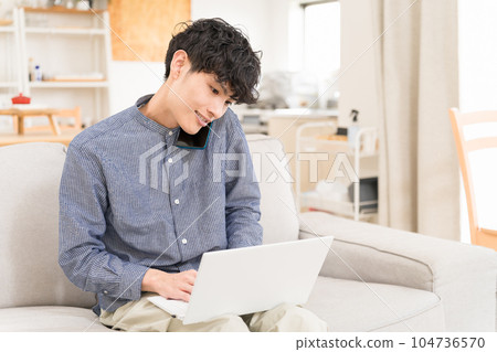 Young man sitting on a sofa with a computer on his lap and calling on a smartphone 104736570