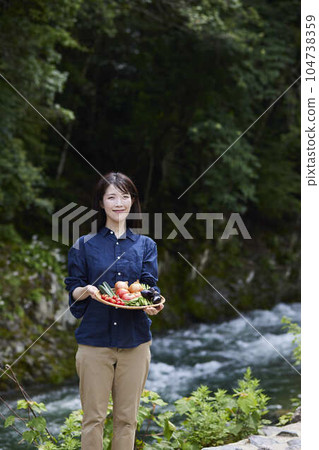 Japanese woman farmer with freshly picked vegetables in front of the river Japanese woman farmer with freshly picked vegetables in front of the river 104738359