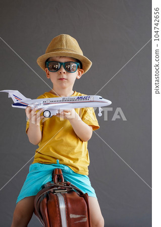 A child in summer clothes and sunglasses sits on a suitcase and holds a toy airplane in his hands. Waiting for travel. Family vacation. Dreams of travel. Summer vacation. 104742656