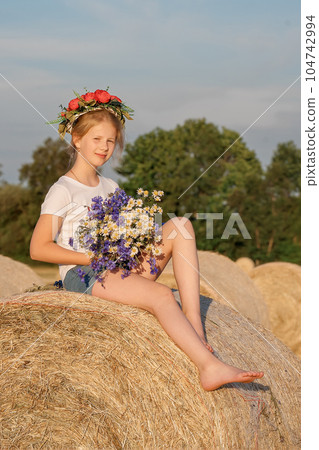 Attractive girl with a wreath and a bouquet of flowers sitting on a haystack. 104742994