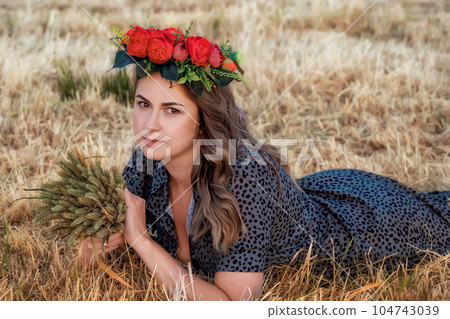 Pretty girl in a summer dress and with a wreath of red roses lying on a summer field. Summer portrait. A Young Beautiful Girl Lying With Flowers Wreath. Pretty girl in a summer dress and with a wreath of red roses lying on a summer field. Summer portrait. A Young Beautiful Girl Lying With Flowers Wreath. 104743039