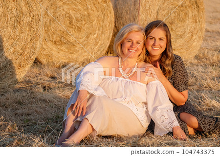 Happy lesbian couple sitting in a field by a haystack. Smiling young lesbian couple. Happy lesbian couple sitting in a field by a haystack. Smiling young lesbian couple. 104743375