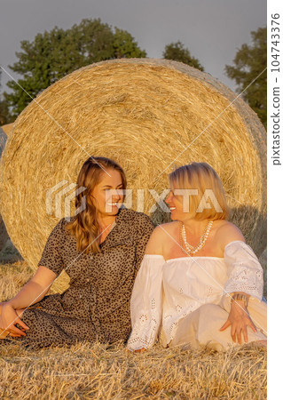 Happy lesbian couple sitting in a field by a haystack and looking at each other with love. Same sex family, happy women. Smiling young lesbian couple. Happy lesbian couple sitting in a field by a haystack and looking at each other with love. Same sex family, happy women. Smiling young lesbian couple. 104743376