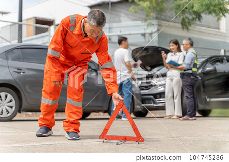 A car insurance agent in orange jumpsuit placing a warning triangle down on the road near the car accident while another insurance agent talking to the car owners in the background A car insurance agent in orange jumpsuit placing a warning triangle down on the road near the car accident while another insurance agent talking to the car owners in the background 104745286