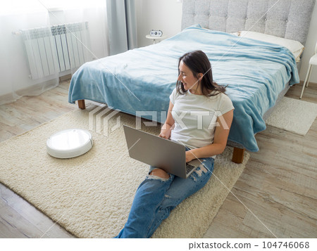 White robotic vacuum cleaner cleaning the floor while woman using laptop 104746068