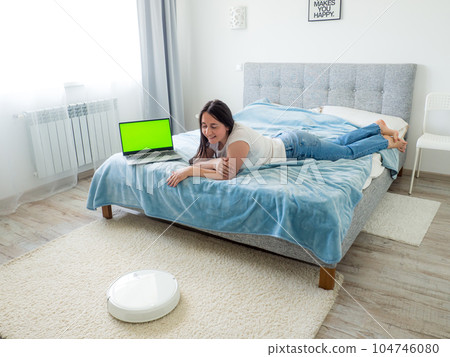 White robotic vacuum cleaner cleaning the floor while woman using laptop White robotic vacuum cleaner cleaning the floor while woman using laptop 104746080