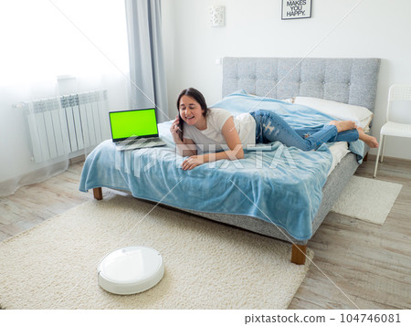 White robotic vacuum cleaner cleaning the floor while woman using laptop 104746081