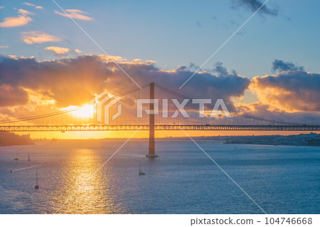 View of 25 de Abril Bridge famous tourist landmark of Lisbon connects Lisboa and Almada on Setubal Peninsula over Tagus river with tourist yacht silhouette at sunset and flying plane. Lisbon, Portugal View of 25 de Abril Bridge famous tourist landmark of Lisbon connects Lisboa and Almada on Setubal Peninsula over Tagus river with tourist yacht silhouette at sunset and flying plane. Lisbon, Portugal 104746668