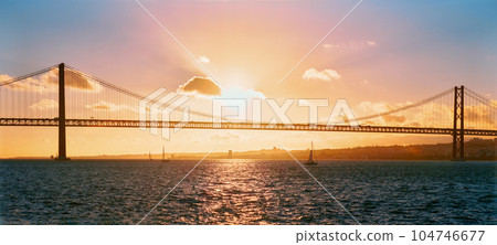 Panorama of 25 de Abril Bridge famous tourist landmark of Lisbon connecting Lisboa and Almada on Setubal Peninsula over Tagus river with tourist yacht silhouette at sunset. Lisbon, Portugal 104746677