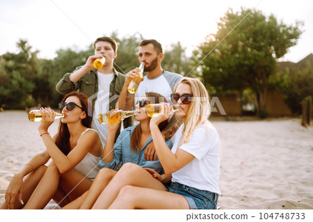 Group of friends cheers and drink beers on the beach. Young friends relaxing and having picnic. Suummertime. Group of friends cheers and drink beers on the beach. Young friends relaxing and having picnic. Suummertime. 104748733