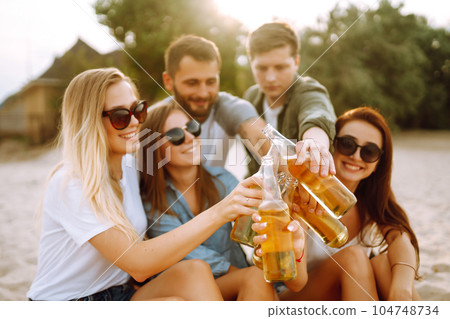 Group of friends cheers and drink beers on the beach. Young friends relaxing and having picnic. Suummertime. Group of friends cheers and drink beers on the beach. Young friends relaxing and having picnic. Suummertime. 104748734