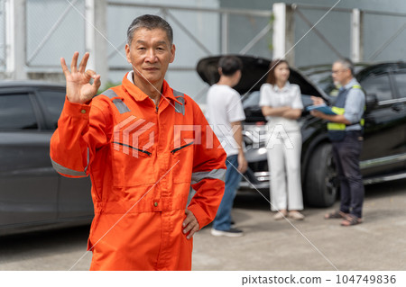 A male car insurance agent in orange jumpsuit standing proudly putting his right hand up with an okay sign and his left hand on his waist, another agent and 2 clients standing in background 104749836