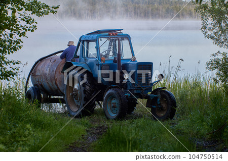 Farmer draws water from lake into tanker attached to wheeled tractor early in morning. 104750514