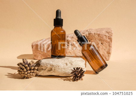 Dark amber glass bottle standing on stone with pine cone. Natural skin care SPA beauty product design. Mineral organic oil cosmetics on beige background. Mock-Up. Oily pipette. Face and body treatment 104751025