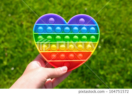 Popit - Colorful antistress sensory toy. Silicone toy - pop it in teen hand on green nature background. Rainbow color, concave and convex bubbles. Trend of summer 2021. Entertainment for children Popit - Colorful antistress sensory toy. Silicone toy - pop it in teen hand on green nature background. Rainbow color, concave and convex bubbles. Trend of summer 2021. Entertainment for children 104751572