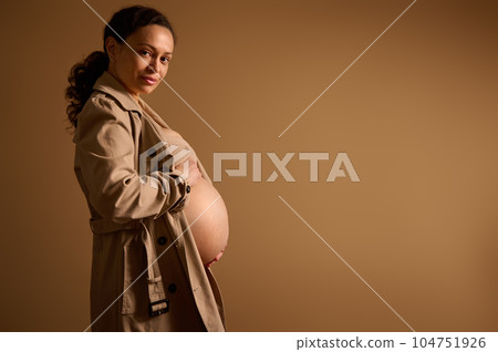 Beautiful pregnant woman in beige trench coat, caressing her big bare belly in late pregnancy, looking at camera Beautiful pregnant woman in beige trench coat, caressing her big bare belly in late pregnancy, looking at camera 104751926
