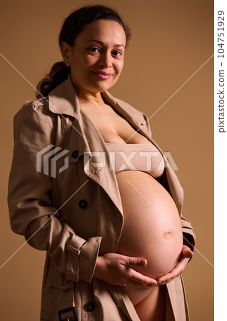 Beautiful pregnant woman in beige trench coat, gently stroking her bare belly, looking at camera, isolated on beige Beautiful pregnant woman in beige trench coat, gently stroking her bare belly, looking at camera, isolated on beige 104751929
