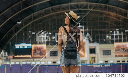 Rear view of young female traveller standing with backpack and hat at train station looking around for sign board to travel towards destination during vacation Rear view of young female traveller standing with backpack and hat at train station looking around for sign board to travel towards destination during vacation 104754502