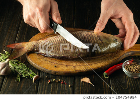 The cook cuts fresh fish on a wooden cutting board with knife. Home cooking of a delicious fish dish according to an old recipe 104754984