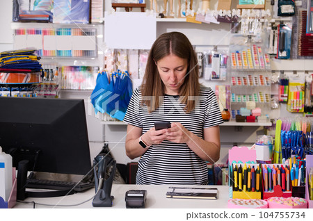 Young woman manager, seller in creative store, using mobile phone, standing at the counter and ordering stationery 104755734