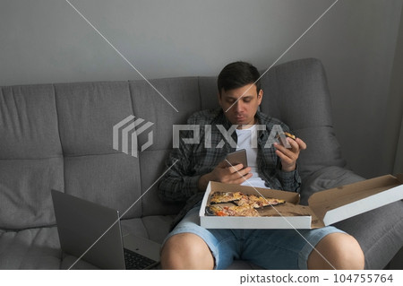 Man bachelor sits on sofa eating tasty pizza, drinking tea, browsing smartphone. 104755764