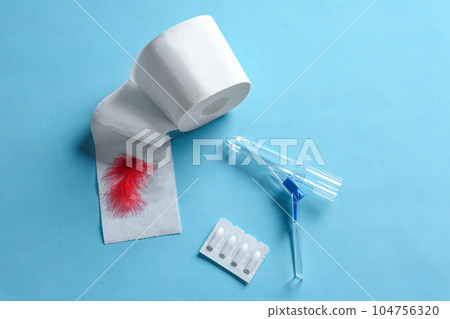 Anoscope, suppositories and toilet paper with red feather on light blue background, flat lay. Hemorrhoid treatment 104756320