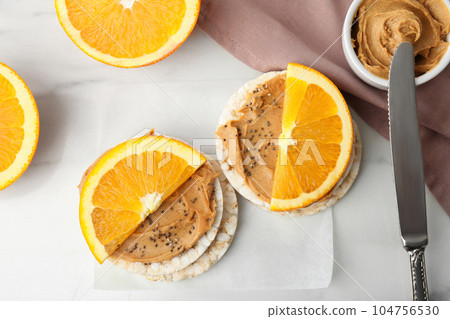 Puffed rice cakes with peanut butter and orange on white marble table, flat lay Puffed rice cakes with peanut butter and orange on white marble table, flat lay 104756530