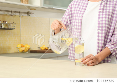 Young woman pouring lemon water into glass from jug in kitchen, closeup. Space for text Young woman pouring lemon water into glass from jug in kitchen, closeup. Space for text 104756789