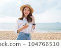 woman wear hat and standing at beach, hand holding smartphone with using social media on internet. 104766510
