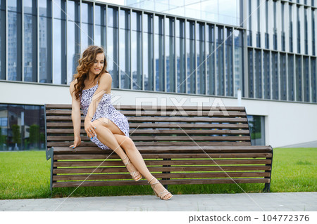 Playful woman in sundress crossed her legs while sitting on bench Playful woman in sundress crossed her legs while sitting on bench 104772376