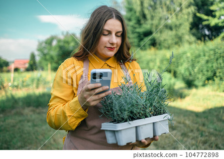 Phone call. Portrait of a young woman carrying flower pot with lavender while taking care of plants in the beautiful garden field. Caucasian farmer using a smartphone Phone call. Portrait of a young woman carrying flower pot with lavender while taking care of plants in the beautiful garden field. Caucasian farmer using a smartphone 104772898