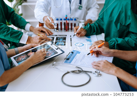 Medical team having a meeting with doctors in white lab coats and surgical scrubs seated at a table discussing a patients working online using computers 104773253