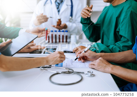 Medical team having a meeting with doctors in white lab coats and surgical scrubs seated at a table discussing a patients working online using computers 104773258