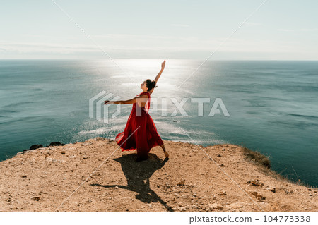 Woman red dress sea. Female dancer posing on a rocky outcrop high above the sea. Girl on the nature on blue sky background. Fashion photo. Woman red dress sea. Female dancer posing on a rocky outcrop high above the sea. Girl on the nature on blue sky background. Fashion photo. 104773338