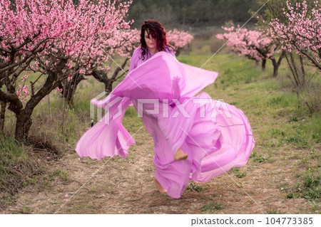 woman blooming peach orchard. A woman in a long pink dress walks in the park, in a peach orchard. Large blooming peach orchard. woman blooming peach orchard. A woman in a long pink dress walks in the park, in a peach orchard. Large blooming peach orchard. 104773385