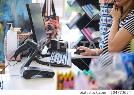 Woman seller in office supply shop, talks on mobile phone, works on PC computer, orders merchandise 104773634