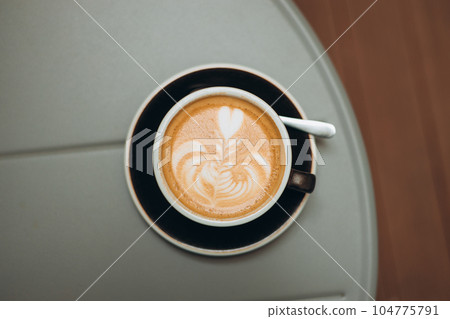 Top view of tasty cappuccino cup stand on steel gray table. One black Cup of cappuccino with latte art. Coffee in ceramic mug in cafe. Concept of easy breakfast. Top view Top view of tasty cappuccino cup stand on steel gray table. One black Cup of cappuccino with latte art. Coffee in ceramic mug in cafe. Concept of easy breakfast. Top view 104775791