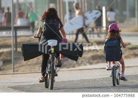 Tourists ride a bike with yoga mats and go to the beach holding a surfboard 104775988