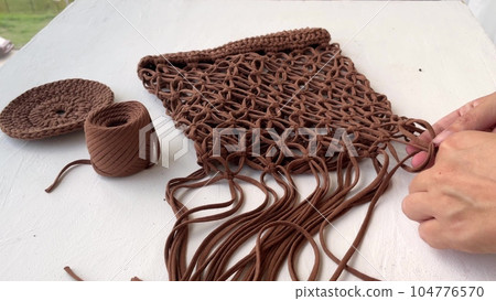 Close-up of women's hands weaving a string bag from a brown knitted cord in the macrame technique. Do it yourself. 104776570