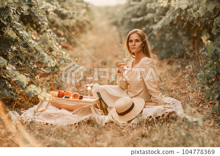 Woman picnic vineyard. Romantic dinner, fruit and wine. Happy woman with a glass of wine at a picnic in the vineyard on sunny day, wine tasting at sunset. 104778369