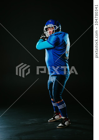 Portrait of a man in a blue uniform for american football throws the ball on a black background.  104780341