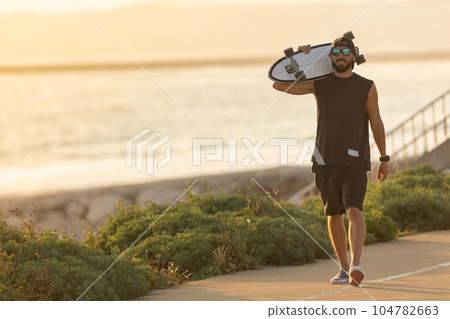 A man walking on the road near by beach holding a skateboard A man walking on the road near by beach holding a skateboard 104782663