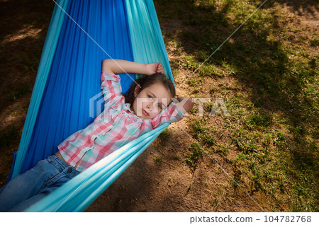Top view lovely little kid girl lying on a blue hammock, relaxing, smiling looking at camera. Happy childhood. Children Top view lovely little kid girl lying on a blue hammock, relaxing, smiling looking at camera. Happy childhood. Children 104782768