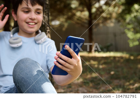 Focus on modern smartphone in hand of a preteen boy talking via video link while resting outdoors. Online communication Focus on modern smartphone in hand of a preteen boy talking via video link while resting outdoors. Online communication 104782791